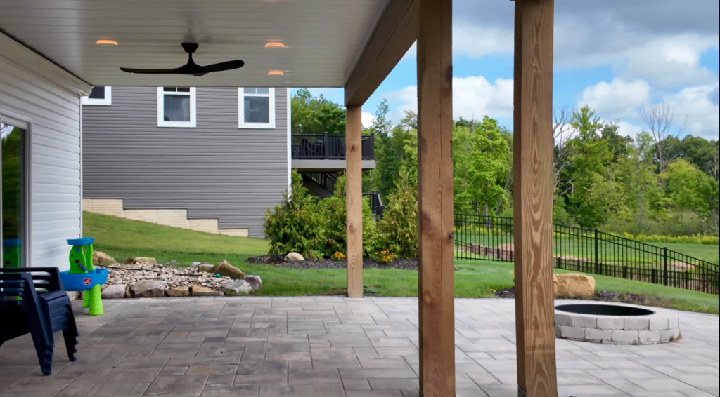 Richfield Two Story Trex Deck With a Cathedral Roof 8 DreamDecks designs and builds architectural-grade outdoor living spaces across Northeast Ohio. From modern decks to full backyard sanctuaries, each project reflects craftsmanship, precision, and timeless luxury. Call 330-907-3040 or visit buildyourDreamDeck.com DreamDecks | Premier Deck Builder & Outdoor Architecture Dry paver patio under the upper deck with a white ceiling, recessed lights, a ceiling fan and wrapped posts, looking out to the fire pit in Richfield Ohio